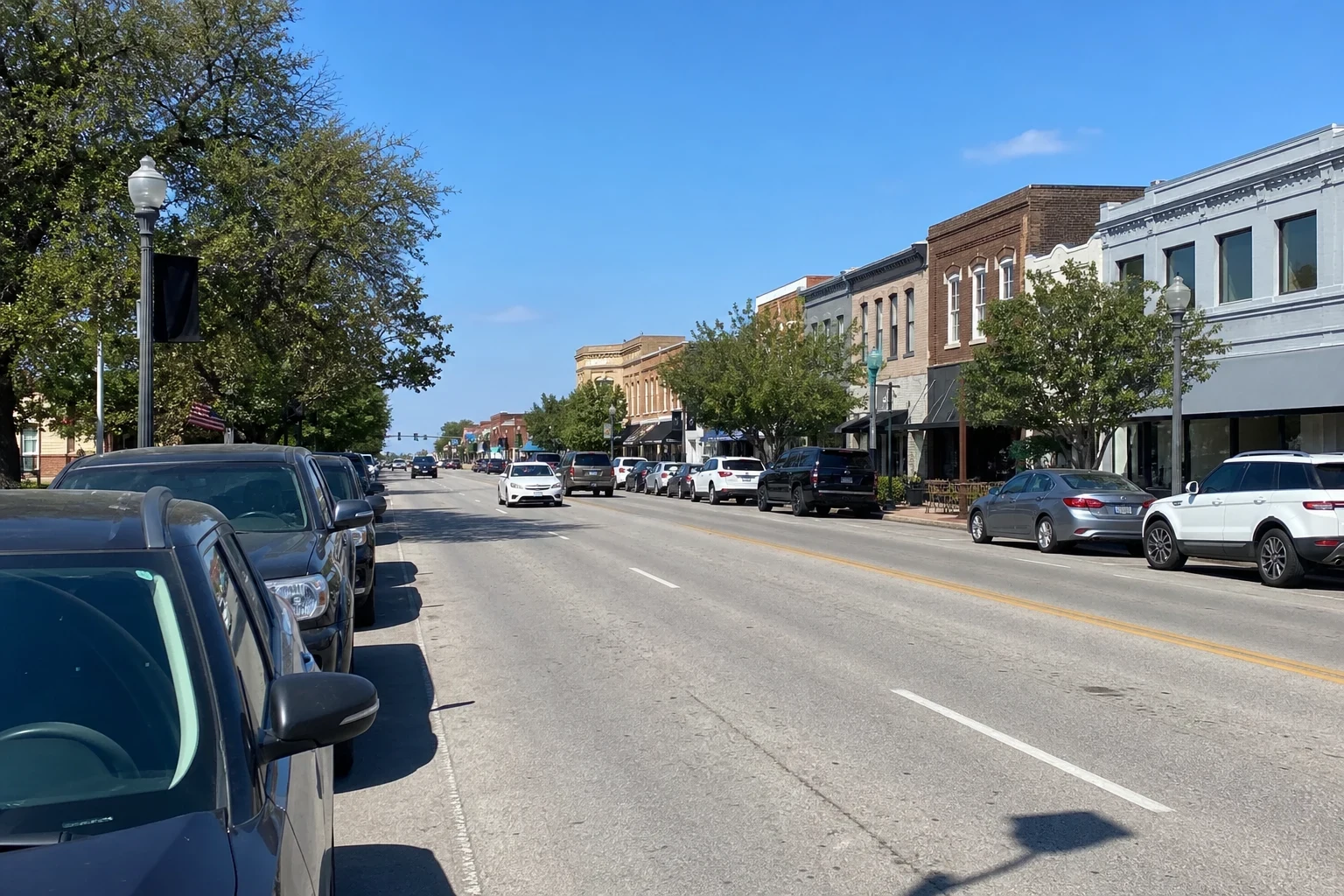 Main street through a small Texas downtown with brick storefronts, street lamps, trees and cars parked along both sides under a clear blue sky