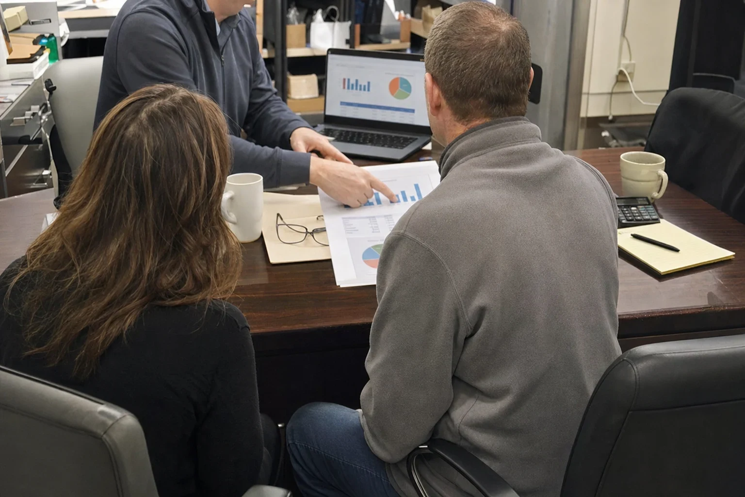 Agent points to a printed chart on a desk while a man and woman, seen from behind, read along in a cluttered back office with shelves, mugs and a laptop